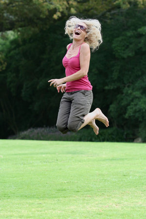 Happy blonde lady in fifties jumping with joy on grass to show excitement and happiness. See my other happy person jumping shots!の写真素材