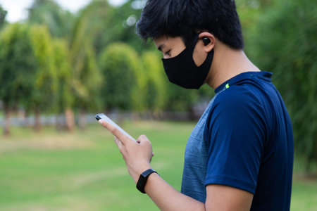 young fitness man with mask at the park wearing wireless earphones and smart watch checking mobile phoneの写真素材