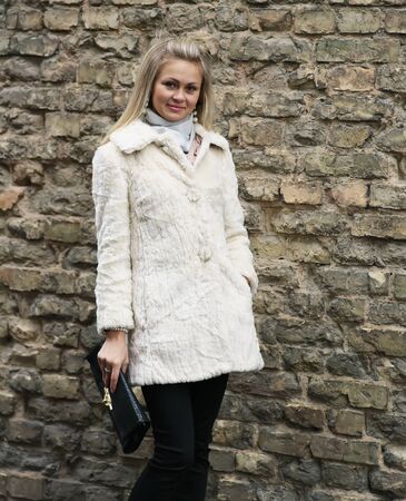 Young Pretty Woman In Fur Coat With Clutch Near Old Brick Wallの写真素材