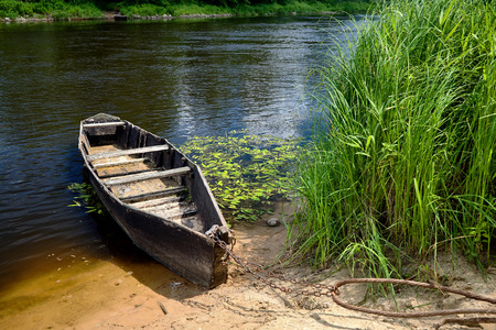Old wooden rowing boat tied with chain and padlock at green summer bank at noon on Neman river in Grodno Belarusの写真素材