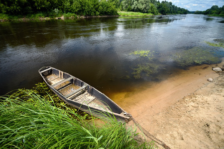 Old wooden rowing boat tied with chain and padlock at green summer bank at noon on Neman river in Grodno Belarus shot wideの写真素材