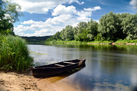 Old wooden rowing fishing boat ferry chained to sandy beach summer afternoon with wind-washed water and grass with ND filterの写真素材