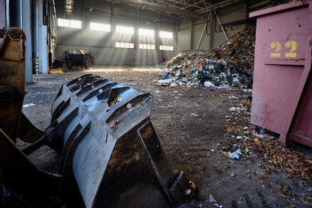 Heavy bulldozer bucket in rays of evening sun in building of waste recycling plant moves tons of garbage into piles for further sorting and processing.のeditorial素材