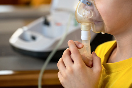Child makes inhalation at home with nebulizer on out of focus background. Example of combating respiratory diseases such as tracheitis bronchitis pneumonia with medical equipment at home conditions.の写真素材