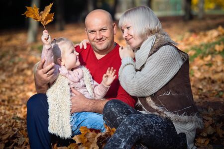 Family idyll during golden autumn. Happy family father mother and little daughter smiling relaxing and having joy in sunny autumn park among fallen golden leaves.の写真素材