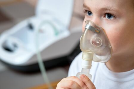 Child makes inhalation at home with nebulizer on out of focus background. Example of combating respiratory diseases such as tracheitis bronchitis pneumonia with medical equipment at home conditions.の写真素材
