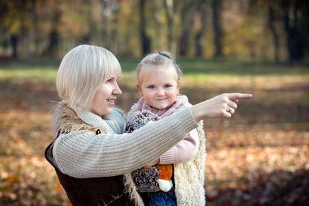 Mom points a finger forward and daughter smiles. Mother and her little child with toy together in sunny park in autumn as happy family.の写真素材