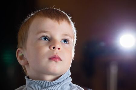 Facial portrait of dreamy pensive blond boy with gaze his eyes fixed upwards over dark background, with specks of dust flying in backlight with flares and copy spaceの写真素材
