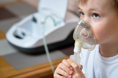 Child makes inhalation at home with nebulizer on out of focus background. Example of combating respiratory diseases such as tracheitis bronchitis pneumonia with medical equipment at home conditions.の写真素材