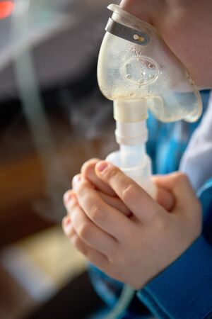 Child makes inhalation at home with nebulizer on out of focus background. Example of combating respiratory diseases such as tracheitis bronchitis pneumonia with medical equipment at home conditions.の写真素材