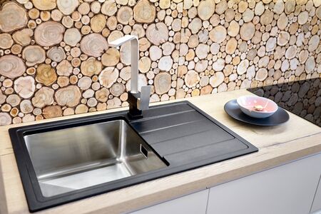 Modern design chrome water tap over stainless steel kitchen sink on table top made of natural wood, with surface behind countertop decorated with wall cross section of tree trunks of cut tree logsの写真素材