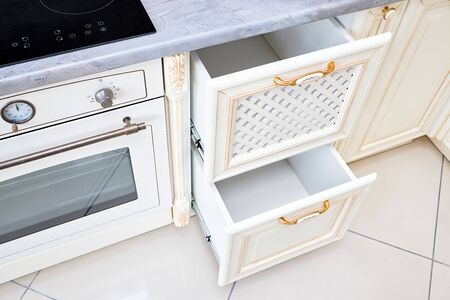 Interior of modern kitchen in classic style with golden elements in beige colors with built-in appliances. Drawer for plates or dishes shelf under the countertop with lattice front panel made of woodの写真素材
