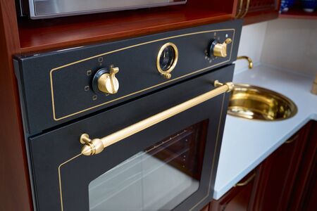 Part of modern kitchen interior in classic style cherry alder wood cabinetry built-in electric oven with golden elements, controls analog timer golden sink with white stone countertop backgroundの写真素材