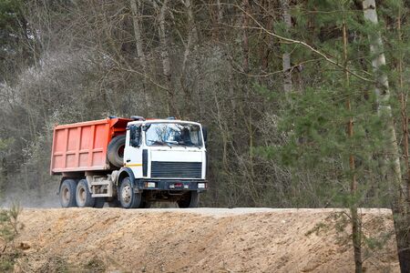 Heavy laden multi-ton truck with white cab and orange body rides along dusty country road in with spring forest background, leaving behind clouds of dust.の写真素材