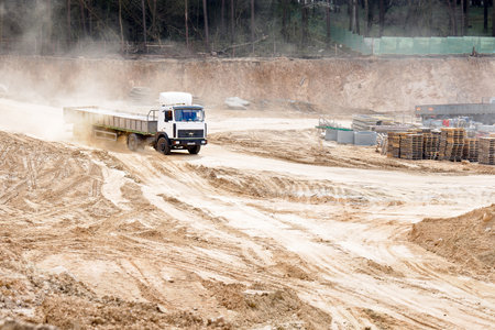GRODNO, BELARUS - APRIL 2020: Heavy laden multi-ton truck MAZ white cab and gray body arrives at the construction site loaded with reinforced concrete near spring forest leaves behind clouds of dust.のeditorial素材