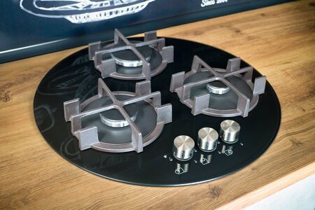 Modern kitchen equipment concept, black tempered glass round shape gas hob with cast-iron grill with three gas burners on brushed oak table top for safe and convenient cooking at home on gas cooktop.の写真素材
