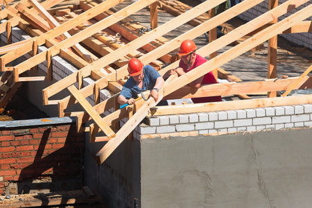 Two workers roofers in helmets surrounded by rafters and beams at construction site build house. Pair of carpenters set up timber roof trussのeditorial素材