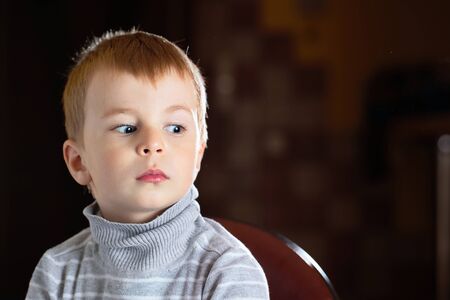 Facial portrait of scared pensive blond boy looking away with fear over dark background with specks of dust flying in backlight with flares and copy space.の写真素材