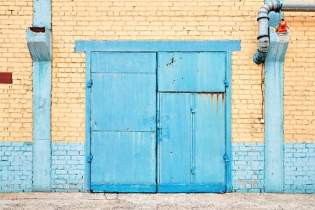 Cityscape. Urban symmetric view of abandoned factory building blue painted rusty gate on yellow brick wall with explosion-proof lantern and copyspace.の写真素材