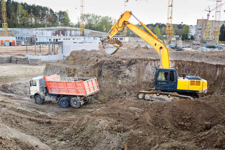 Crawler excavator in process of loading sand soil to multi-ton mining truck at construction site with cranes, buildings and workers in background.の写真素材