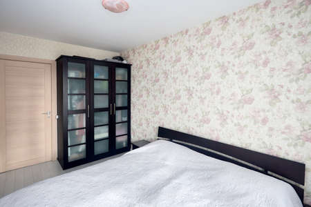 Small bedroom with simple bed with bedspread and flowered walls and curtains balcony or loggia. Concept of real bedroom of real estate or hotel.の写真素材