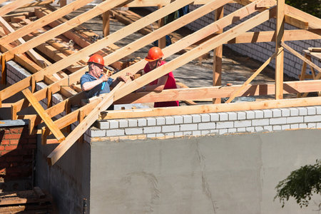 GRODNO, BELARUS - JUNE 2020: Two workers roofers in helmets surrounded by rafters and beams at construction site build house. Pair of carpenters set up timber roof truss, knock with hammer hold board.のeditorial素材