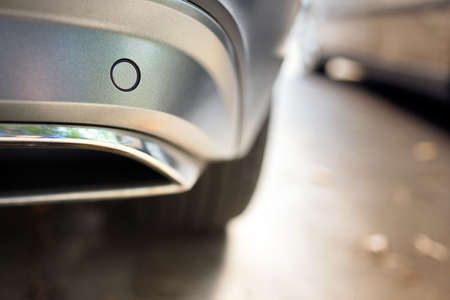 Selective focus on rear bumper parking sensor near exhaust pipe at back of luxurious car bumper close up near other car on light blurred with sun flare background with copyspace.の写真素材