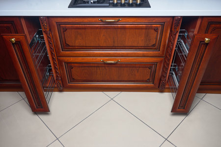 Pull out spice rack cabinet filler pantry. Two vertical spices drawers shelf under countertop with wooden panel and hob. Classic style kitchen with golden elements, cherry alder wood cabinetry.の写真素材
