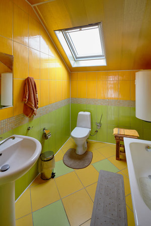 Interior of modern bathroom and toilet in attic room. Bath, toilet, trash can, hot water boiler mirror, dormer window, yellow and green ceramic tiles on the floor and walls.の写真素材