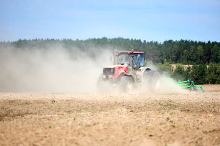Tractor is plowing field with harrow. Typical agricultural scene tractor cultivation in field in clouds of dust. Selective focus red farm tractor with plow spring field under blue sky with copyspace.の写真素材