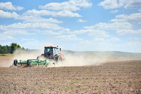 Typical agricultural scene tractor cultivation in field in clouds of dust drives off into the distance. Selective focus red farm tractor with green plow spring field under blue sky with copyspace.の写真素材