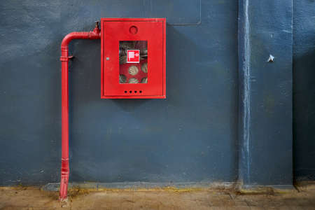 Fire hose in red box. Pipe roll for fire emergency in red metal cabinet on gray painted concrete wall with tiled floor as part of firefighting system of industrial production plant with copyspace.の写真素材