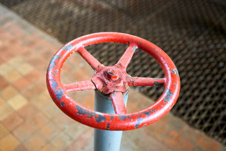 Old rusty painted red valve wheel with swellings and peeling selective focus with extension device over out of focus background tiled floor and perforated metal sheet stamping plates centered.の写真素材