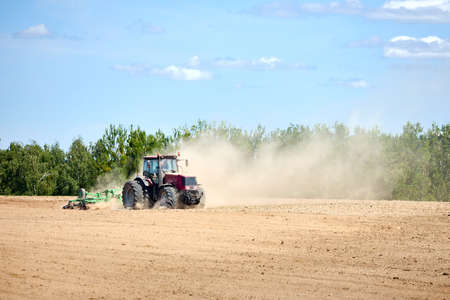 Tractor is plowing field with harrow. Typical agricultural scene tractor cultivation in field in clouds of dust. Selective focus red farm tractor with plow spring field under blue sky with copyspace.の写真素材