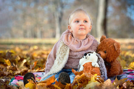 Little beautiful dreamy caucasian girl with earrings in autumn park sits in golden autumn leaves with with soft toys hedgehog and bear cub over fall background with copyspace.の写真素材