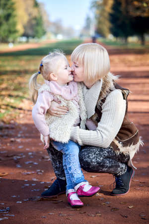 Scene of quiet family happiness and love. Happy caucasian mother holding her little daughter on her laps face to face closely in park together in warm sunny autumn day, vertical shot.の写真素材