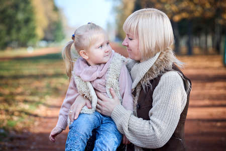 Scene of quiet family happiness filiation and love. Happy caucasian mother holding her little daughter on her laps face to face closely looking into each others eyes in park in warm sunny autumn day.の写真素材
