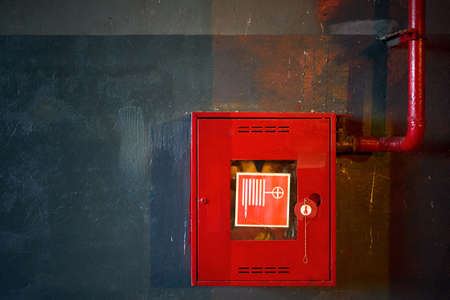 Fire hose in red box. Pipe roll for fire emergency in red metal cabinet on grey oil paint concrete wall with multicolor reflections as part of fire fighting system of industrial production plant.の写真素材