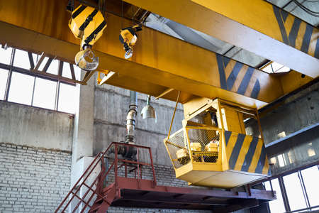 Yellow with black stripes overhead crane with linear traverse and hooks at petrochemical enterprises chemical plant workshop. Crane operator cabin and hooks and jib crab trolley. Landing staircase.の写真素材