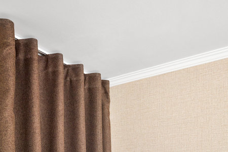 Curtains made of brown matting fabric hang on the ceiling cornice near the wall papered with a matting texture or rough canvas under a white plasterboard ceiling with ceiling baseboards.の写真素材