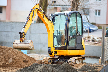 Tracked orange mini excavator digger near road with green trees and concrete fence on background with copyspace.の写真素材
