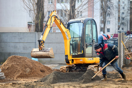 Grodno, Belarus - November, 22 2021: JCB 8018 CTS excavator digging sand and concrete at construction site with construction workers with shovels. Concept of collaboration of machines and people.のeditorial素材