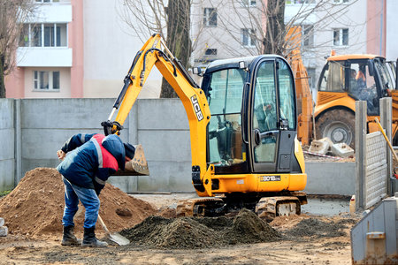 Grodno, Belarus - November, 22 2021: JCB 8018 CTS excavator digging sand and concrete at construction site with construction workers with shovels. Concept of collaboration of machines and people.のeditorial素材