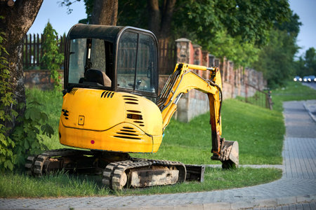 Tracked orange mini excavator digger near road with green trees and stone fence and cars lights on background with copyspace.の写真素材