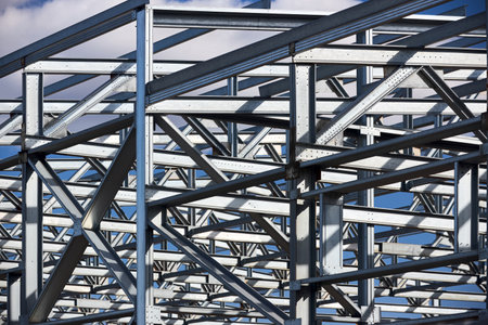 Metal construction. Urban geometrical interlacing of bolted connections cross bars and beams of construction site over blue sky with clouds background.の写真素材