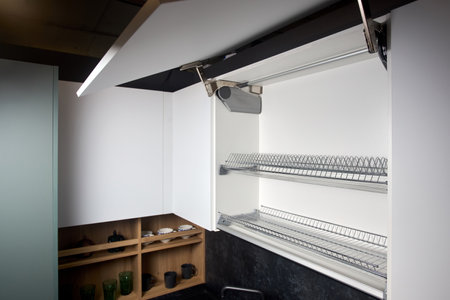 System for bi-fold fronts and facades for kitchen cabinets direct access with lifting mechanism closeup. Fitted flat design kitchen with sink tap and opened cupboard sideboard dish drying cabinet.の写真素材