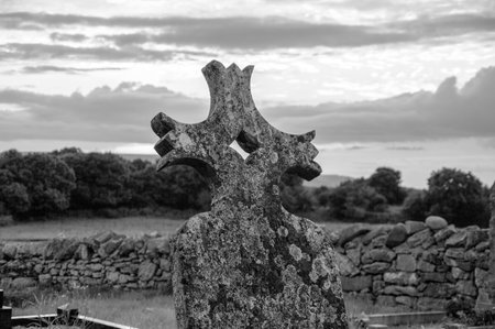 A stone cross is standing in a field with a wall in the background. The sky is cloudy and the sun is settingの写真素材