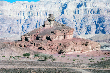 A rocky mountain with a large rock on top. The mountain is surrounded by a desert. The sky is clear...