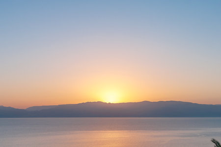 The sky is a beautiful shade of blue with a bright orange sun in the background. The water is calm and peaceful, and the mountains in the distance add to the serene atmosphereの写真素材