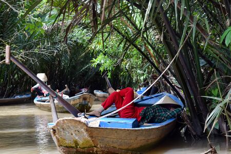 Oarsman resting in the boat in delta of Mekong river, Vietnam. Bright red trousers provide great contrast in the wilds of the green jungleの写真素材